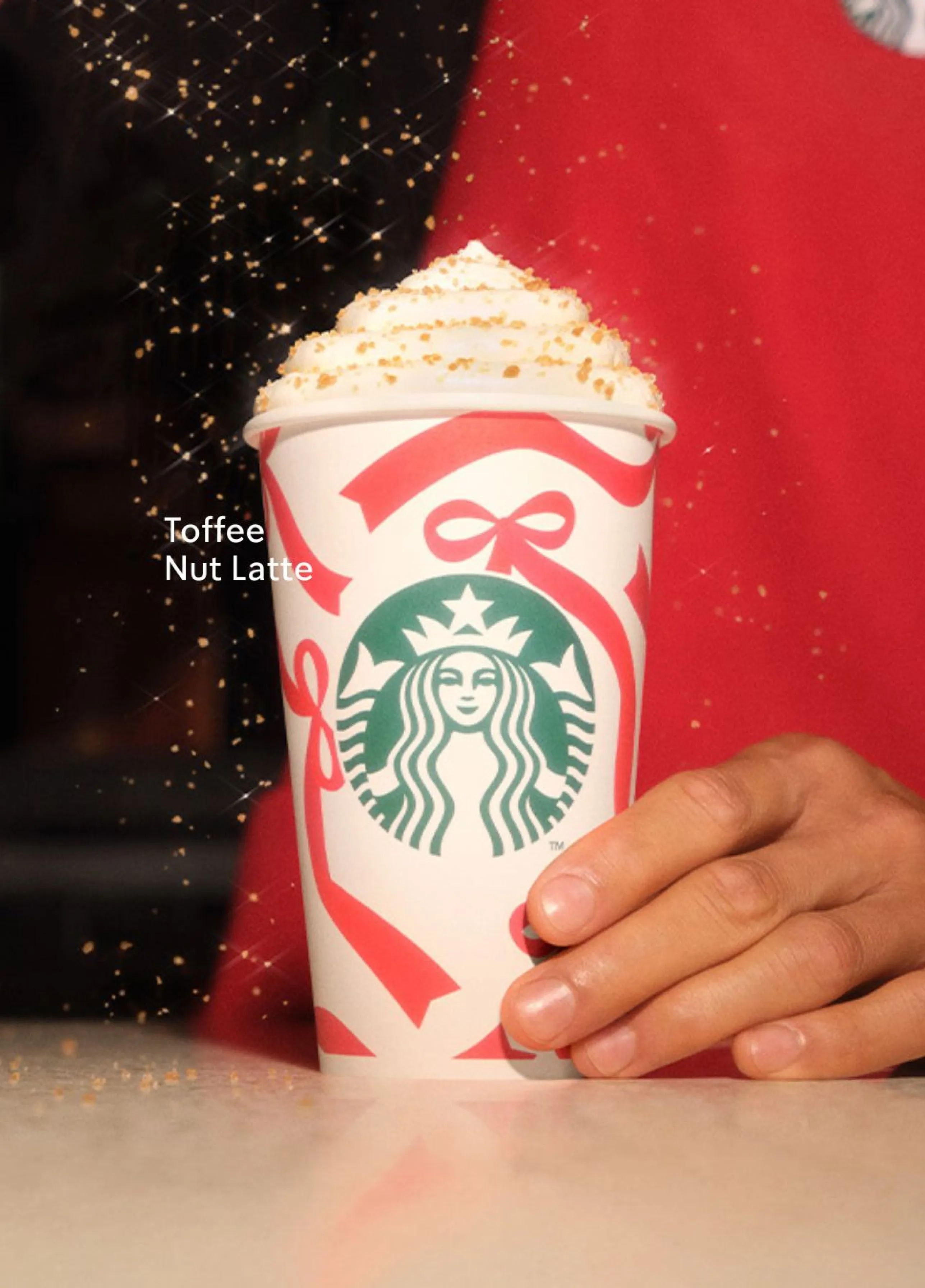 Kubek Starbucks w świątecznym opakowaniu z czerwonymi wstążkami stoi na blacie. Na wierzchu napoju znajduje się bita śmietana posypana karmelową posypką. Obok kubka widoczna jest dłoń osoby w czerwonym fartuchu, która dodaje posypkę. Na kubku widnieje logo Starbucks, a obok napis „Toffee Nut Latte”. W tle znajdują się pionowe linie, sugerujące wnętrze kawiarni.