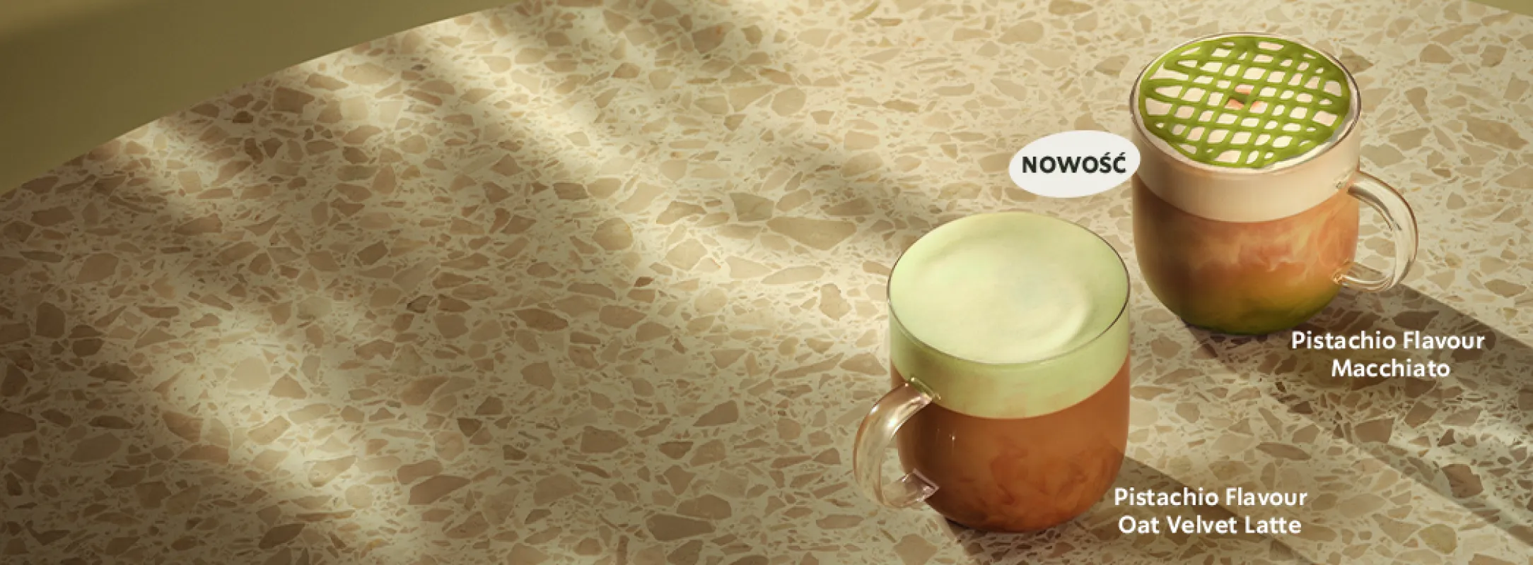 Dwa szklane kubki z kawami o smaku pistacjowym stoją na jasnym, kamiennym blacie w ciepłym świetle. Pierwszy napój to Pistachio Flavour Oat Velvet Latte z jasnozieloną pianką, drugi to Pistachio Flavour Macchiato z dekoracją w formie zielonej kratki na wierzchu. Obok napoju Macchiato znajduje się etykieta z napisem ‘NOWOŚĆ’