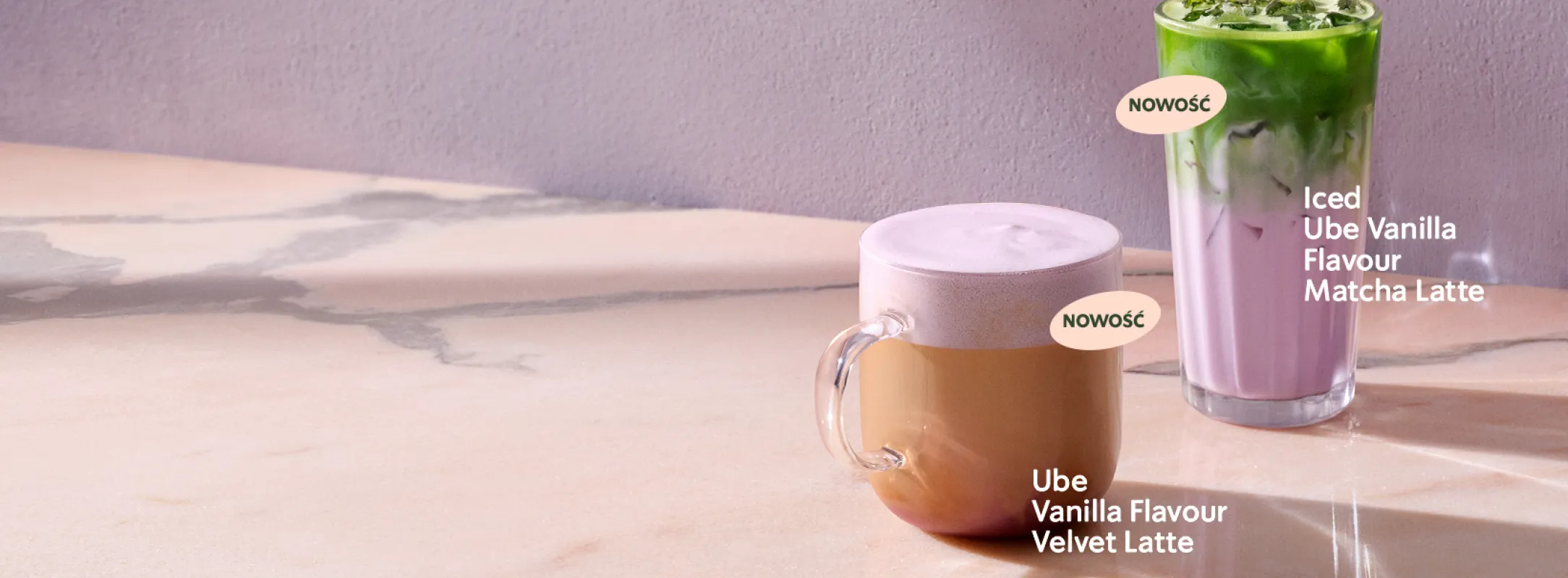 "Na jasnym, marmurowym blacie stoją dwa napoje Starbucks. Po lewej znajduje się filiżanka z fioletową pianką opisana jako ‘Nowość – Ube Vanilla Flavour Velvet Latte’. Po prawej stoi wysoka szklanka z warstwowym napojem: fioletowym mlekiem i zieloną herbatą matcha, opisana jako ‘Nowość – Iced Ube Vanilla Flavour Matcha Latte’. Tło jest jasnofioletowe.